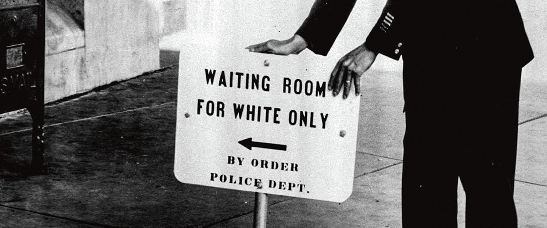 Black and white photo of a person placing down a sign that says "Waiting Room for White Only"