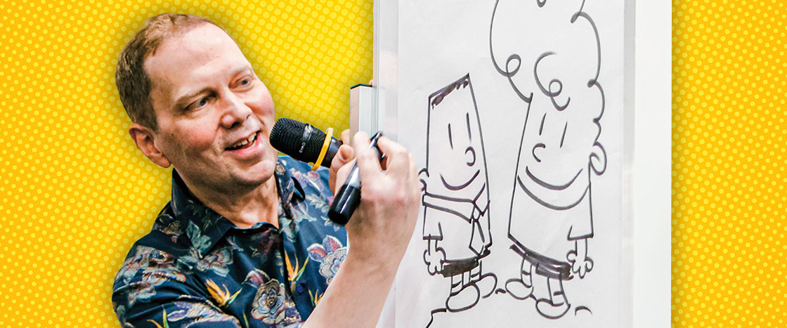 A man drawing two boys on a white board as he speaks into a microphone